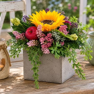 Puede incluir: Una maceta cuadrada de hormigón gris con un arreglo floral que incluye un gran girasol amarillo, una manzana roja, racimos de pequeñas flores rosas, ramitas moradas y follaje verde. El arreglo es denso y tiene mucha textura.