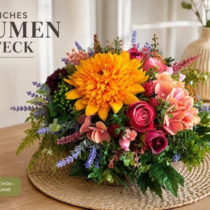 Pode incluir: Um arranjo floral com uma grande dália laranja central, rosas rosa-escuras, cachos de hortênsias rosa-claras, ramos de lavanda roxa e folhagem verde. O texto KÜNSTLICHES BLUMEN GESTECK, PFLEGELEICHT, LANGLEBIG, VIELSEITIGE DEKO e NATÜRLICH SCHÖN – DAS GANZE JAHR é visível.
