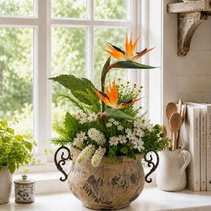 Könnte beinhalten: Ein dekoratives Arrangement aus künstlichen Paradiesvogelblumen mit orangefarbenen und blauen Blütenblättern, weißen Blüten und grünem Blattwerk. Die Blumen befinden sich in einem runden, verwitterten beigen Topf mit zwei schwarzen Metallhenkeln.