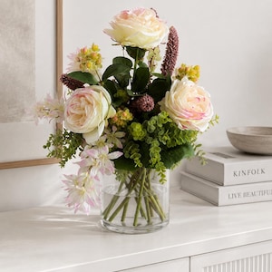 Könnte beinhalten: Ein Blumenarrangement in einer klaren Glasvase mit cremefarbenen und blassrosa Rosen, kleinen grünen Beeren, violetten ährenartigen Blüten und zarten hellrosa Blüten mit grünem Blattwerk. Die Stiele sind durch das Glas hindurch sichtbar.