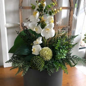 Op de afbeelding: Een bloemstuk in een matte donkergrijze cilindrische pot met witte orchideeën, groen varenachtig blad, twee ronde gestructureerde groene bloemhoofdjes en dunne bruine decoratieve takjes voor een houten raamkozijn.