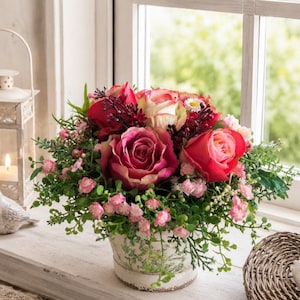 Op de afbeelding: Een bloemstuk met donkerroze en crèmekleurige rozen, kleine roze bloesems en groen blad. De bloemen zitten in een ronde, gebroken witte keramische pot, versierd met een subtiel groen botanisch patroon en een rand aan de onderzijde.