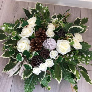 May include: A floral arrangement featuring white roses, green and white variegated leaves, brown pinecones, dark brown lotus pods, and small white flowers. A light-coloured ribbon with the printed text In Erinnerung is tucked into the foliage.