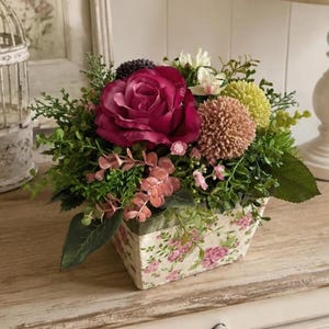 Può includere: Una composizione floreale in un contenitore rettangolare con motivo a rose rosa. Il bouquet presenta una grande rosa magenta scuro, un fiore a pompon testurizzato color pesca, un fiore sferico verde chiaro e vari fogliami verdi e piccoli fiori rosa.