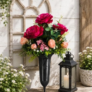 May include: A black plastic cemetery vase with a pointed stake base, filled with an arrangement of deep magenta roses, soft peach-coloured smaller roses, tiny pink filler flowers, and green foliage, positioned upright on a wooden surface.