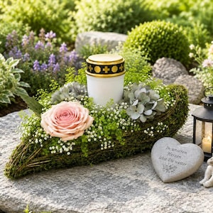 Könnte beinhalten: Eine weiße Grabkerze mit goldenem Deckel und schwarzen Kreuzsymbolen, eingebettet in ein moosbedecktes Gesteck mit einer rosa Rose, weißem Schleierkraut und grauem Laub, ergänzt durch einen herzförmigen Stein und eine kleine Laterne.