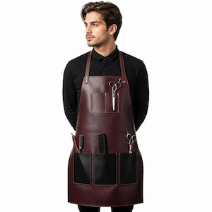 May include: A dark burgundy leather apron featuring a top chest pocket and three lower pockets. The apron includes metal hardware, with silver-toned hair styling scissors and a black comb stored in the pockets for professional utility.