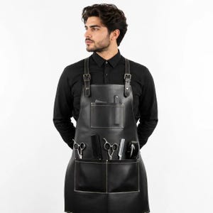 May include: A black leather apron featuring adjustable shoulder straps with metal buckles, a single upper chest pocket, and two lower pockets. The apron contains a black comb, two pairs of silver metal scissors, and a silver metal razor tool.