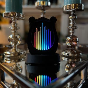 Op de afbeelding: Een zwarte, schildvormige decoratieve lamp met een reeks verticale, verlichte staven die een levendig regenboogverloop weergeven. De lamp is gemonteerd op een stevige zwarte voet en straalt een heldere, veelkleurige gloed uit.