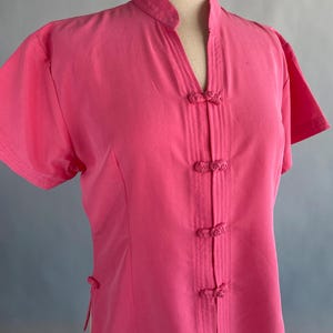Puede incluir: Una camisa rosa brillante de manga corta con cuello mandarín y escote en V. La camisa presenta una serie de cierres decorativos de nudos en la parte delantera y en el lateral. La camisa se exhibe en un maniquí.