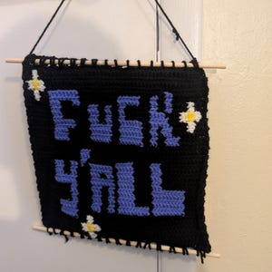 Könnte beinhalten: Ein schwarzer gehäkelter Wandbehang mit dem Text FUCK Y'ALL aus hellblauem Garn. Das Stück ist mit drei kleinen weißen und gelben Blumenmotiven verziert und an zwei hellen Holzstäben mit einer schwarzen Garnaufhängung befestigt.
