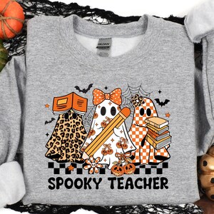 May include: A heather gray crewneck sweatshirt featuring a graphic of three ghosts. One ghost has a leopard print pattern and holds a book, one wears a polka dot bow and holds a pencil, and one has an orange checkerboard pattern with a stack of books. The text reads SPOOKY TEACHER.