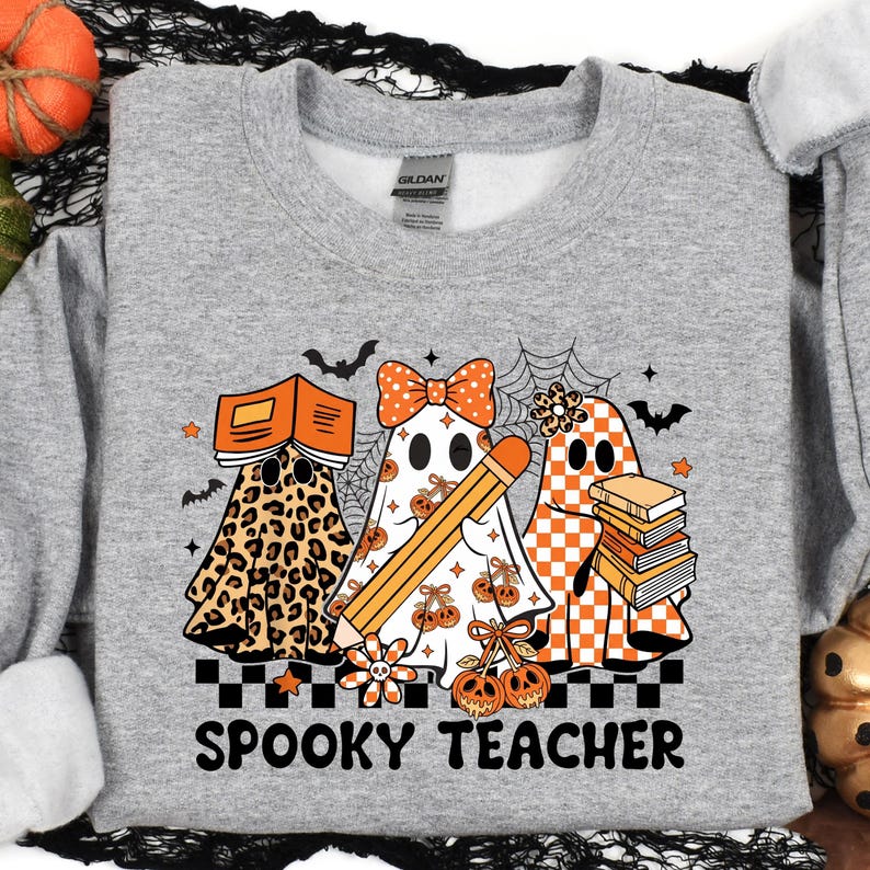 May include: A heather gray crewneck sweatshirt featuring a graphic of three ghosts. One ghost has a leopard print pattern and holds a book, one wears a polka dot bow and holds a pencil, and one has an orange checkerboard pattern with a stack of books. The text reads SPOOKY TEACHER.