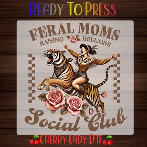 May include: A graphic design featuring a tiger with a person riding on its back, surrounded by pink roses. The text reads READY TO PRESS, FERAL MOMS RAISING HELLIONS, SOCIAL CLUB, and CHERRY LADY DTF. The design uses brown, pink, and orange tones.