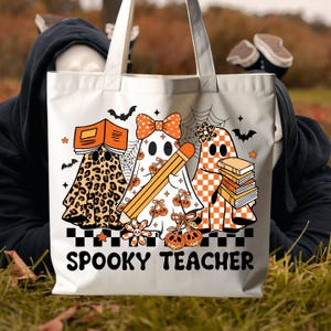 May include: A white tote bag featuring a graphic of three ghosts with orange and black patterns, including leopard print and checkers. The ghosts hold books and a pencil, surrounded by bats and spiderwebs. The text SPOOKY TEACHER is printed below.