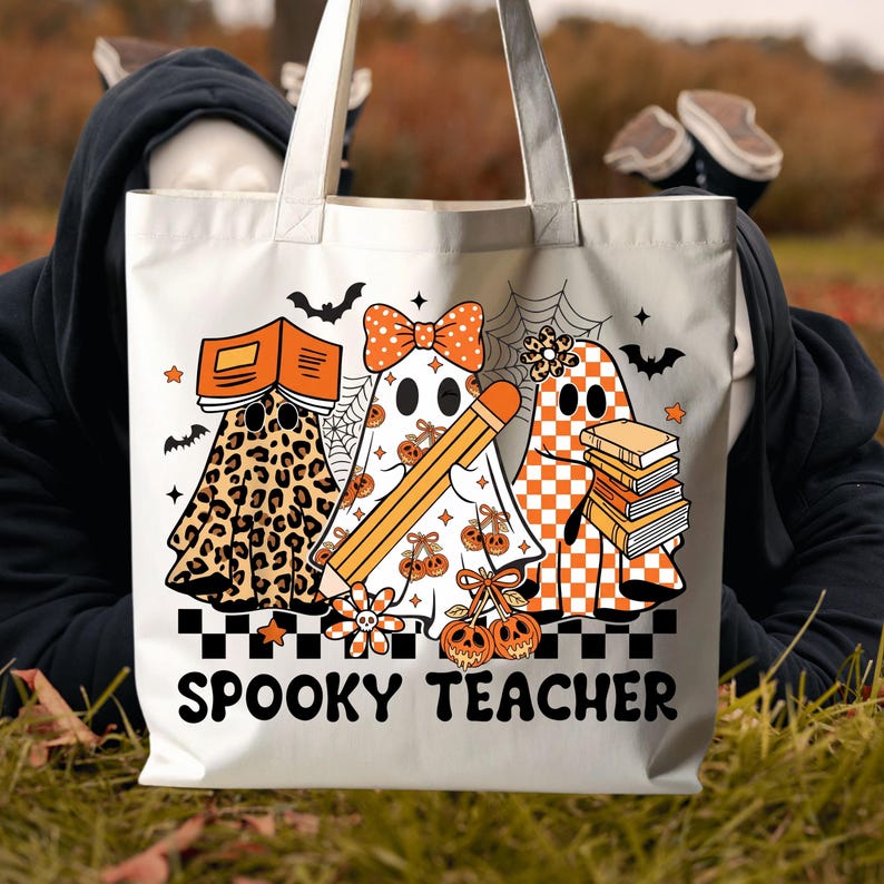 May include: A white tote bag featuring a graphic of three ghosts with orange and black patterns, including leopard print and checkers. The ghosts hold books and a pencil, surrounded by bats and spiderwebs. The text SPOOKY TEACHER is printed below.