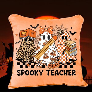 May include: A square orange throw pillow featuring a graphic of three ghosts. One ghost has a leopard print, one has a bow and pencil, and one has a checkered pattern with books. The text SPOOKY TEACHER is printed in black at the bottom.