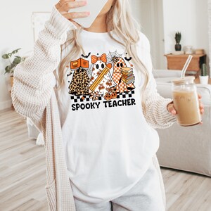 May include: A white t-shirt featuring a graphic of three ghosts with orange and black patterns, including leopard print and checkerboard. The ghosts hold books and a pencil, surrounded by spiderwebs and bats. The text reads SPOOKY TEACHER.