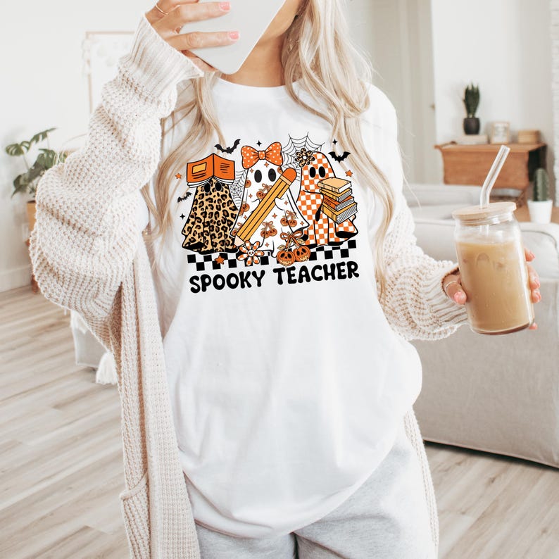 May include: A white t-shirt featuring a graphic of three ghosts with orange and black patterns, including leopard print and checkerboard. The ghosts hold books and a pencil, surrounded by spiderwebs and bats. The text reads SPOOKY TEACHER.
