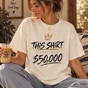 May include: An off-white short-sleeve t-shirt featuring a gold crown graphic above the black text that reads "THIS SHIRT COST ME $50,000" in a stylized, brush-stroke font. The design is centered on the chest of the crew neck garment.