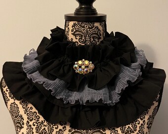 May include: A black and gray ruffled collar with a jeweled brooch. The collar is layered with ruffles and has a delicate, feminine design.