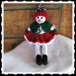 May include: A crocheted snowman doll wearing a red and white hat with a green cape and a red skirt. The doll is sitting on a white surface.