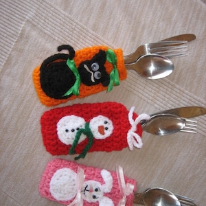 May include: Three crocheted utensil holders, each featuring a different animal design. The first holder is orange with a black cat, the second is red with a snowman, and the third is pink with a white bunny.