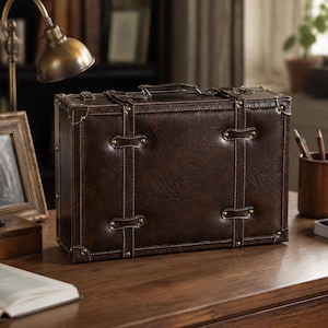 May include: A rectangular dark brown suitcase with a textured finish, featuring two vertical straps with brass-coloured studs, reinforced corner caps, and a top handle. The design includes white contrast stitching along the edges and straps.