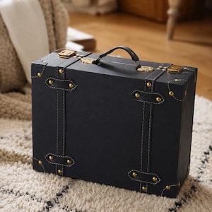 May include: A rectangular black suitcase featuring a top handle, two gold-toned metal latches, and decorative black straps with gold-toned metal rivets at the corners and along the front panel. The exterior has a smooth, matte finish.