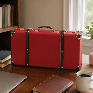 May include: A rectangular red suitcase with dark brown leather-like straps and trim. The case features gold-toned metal corner protectors, buckles, and a top handle. The design includes decorative gold studs along the vertical brown straps.
