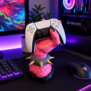 May include: A pink, textured, claw-like hand sculpture with black fingernails holding a white and black wireless game controller. The sculpture features a grey wristband with gold-colored spikes and a black base, designed as a gaming accessory stand.