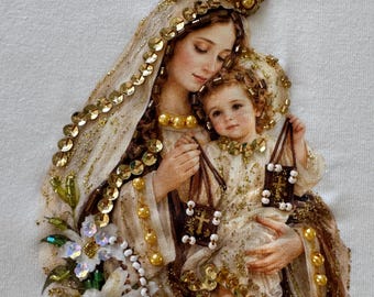 May include: An embroidered fabric appliqué featuring a depiction of a mother and child adorned with gold sequins, beads, and pearls. The figures wear crowns and hold small brown rectangular charms with cross motifs, accented by white floral details.