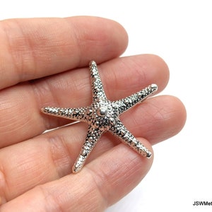 Puede incluir: Un amuleto de estrella de mar de color plateado con una superficie texturizada. La estrella de mar mide aproximadamente 5 cm de largo.
