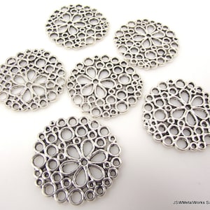 May include: Six silver-coloured, circular metal charms with a floral design in the centre and a pattern of small circles around the edge. These charms are likely used for jewellery making or other craft projects.