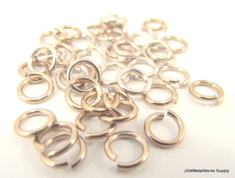 May include: A pile of gold-toned jump rings, also known as split rings, are scattered on a white surface. These rings are used for jewelry making and crafting.