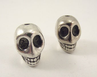 May include: Two silver skull beads with a hole at the top. The skulls have detailed features, including eye sockets, a nose, and a toothy grin.
