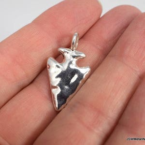 May include: A silver arrowhead pendant with a textured surface. The pendant is shown on a hand.