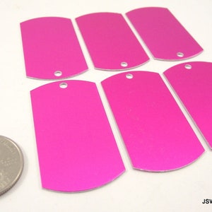 May include: Six blank, pink, metal dog tags with rounded edges and a hole at the top. The tags are about the size of a US quarter.