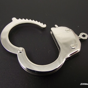 Puede incluir: Esposas de metal plateadas con un diseño de bisagra y un pequeño lazo en un lado. Las esposas están cerradas y descansan sobre una superficie negra.