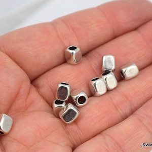 May include: A close-up of silver metal square beads with a hole in the center. The beads are in a hand, ready to be used for jewelry making.