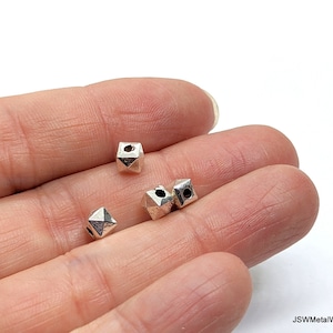 Op de afbeelding: Close-up van verschillende kleine, zilverkleurige geometrische kralen. Elke kraal heeft een veelzijdige, kubusachtige vorm met een gat dat door één kant is geboord. De kralen liggen op een lichtgekleurd oppervlak.