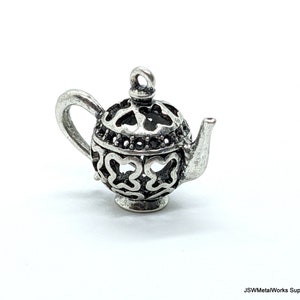 May include: A silver-toned metal teapot charm featuring an intricate openwork cutout design with butterfly-like shapes on the body and lid, a curved handle, a small spout, and a circular loop at the top for attachment. The text JSWMetalWorks Supply is visible.