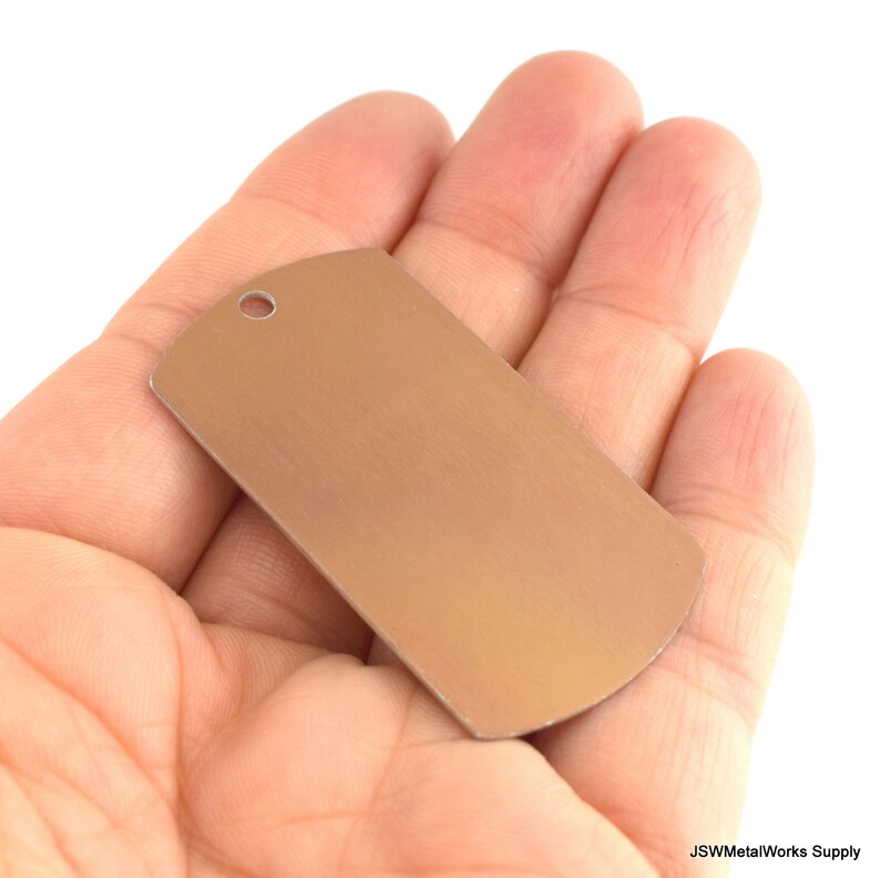 May include: A blank, silver-colored metal dog tag with a rounded top and a hole at the top for a ring or chain. The tag is being held in a hand.