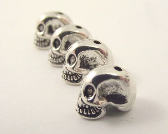 May include: Four silver-colored skull beads arranged in a diagonal line. Each bead has detailed facial features, including eye sockets, a nose cavity, and a grinning mouth. The beads are likely for jewelry making or other crafts.