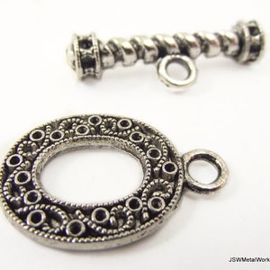 Op de afbeelding: Twee zilverkleurige metalen sieradssluitingen. Eén is een cirkelvormige tuimelsluiting met een sierlijk, opengewerkt ontwerp. De andere is een staafsluiting met een gedraaid touwontwerp. Beide sluitingen hebben kleine lussen voor bevestiging.