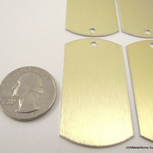 May include: Four blank, gold-colored metal dog tags with rounded edges and a hole at the top. The tags are slightly larger than a US quarter.