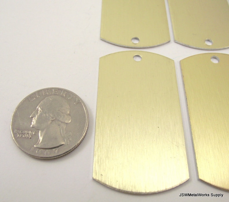 May include: Four blank, gold-colored metal dog tags with rounded edges and a hole at the top. The tags are slightly larger than a US quarter.