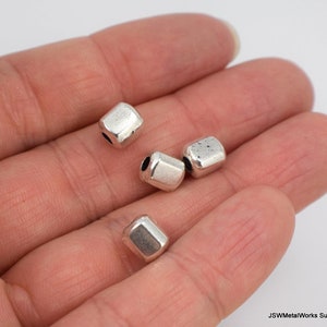 May include: Close-up of several silver-colored, geometric beads. Each bead has a slightly irregular, faceted shape with a hole running through the center. The beads are displayed in a person's hand, showcasing their size and texture. The beads appear to be made of metal.