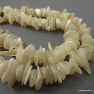 Puede incluir: Una hebra de cuentas de concha blanca, cada cuenta tiene una forma irregular única, con un ligero brillo iridiscente.