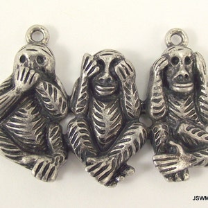 May include: Three silver monkey charms, each with a different gesture: one covering its mouth, one covering its eyes, and one covering its ears. The monkeys are sitting side-by-side.